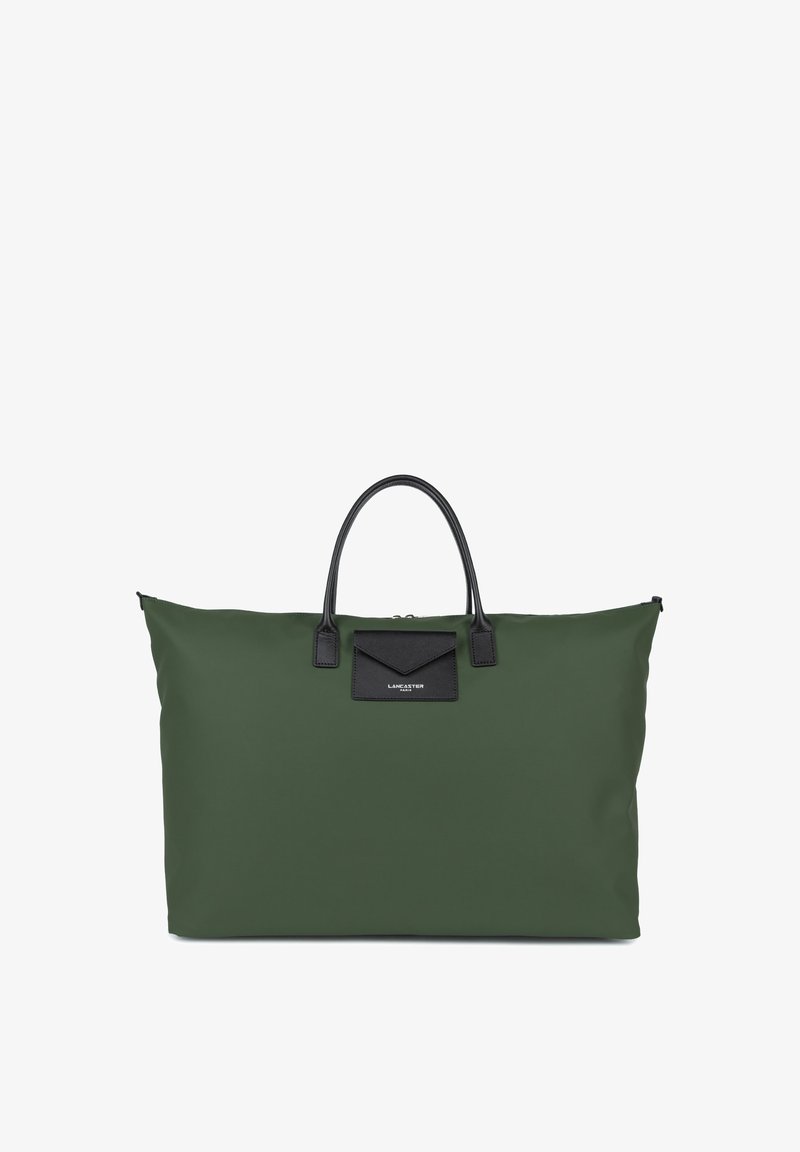Green fabric tote bag with black leather accents, structured shape, dual handles, and a black patch with logo detail on the front.