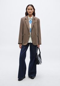 Καφέ υπερμεγέθη σακάκι με χρυσά κουμπιά, ανοιχτό μπλε πουλόβερ αργκλάι, λευκό πουκάμισο, σκούρα τζιν με φουσκωτά μπατζάκια, μαύρες μπότες, μαύρη τσάντα.