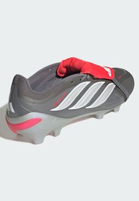 Szürke focicipő piros pánttal, fehér részletekkel és texturált felsőrésszel, kidolgozott talppal és jellegzetes márkajelzéssel az oldalon.