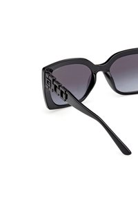 Schwarze Sonnenbrille mit Katzenaugenform, verlaufenden Gläsern und einem geometrischen Akzent am Bügel. Glatte, glänzende Oberfläche.