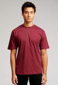 Ung mand med kort mørkt hår iført en ensfarvet maroon T-shirt med kortærmer og sorte bukser, stående mod en neutral baggrund.