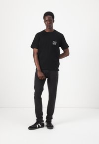 Camiseta de algodón negra con el logo blanco de "GAP" en el pecho, combinada con jeans negros y zapatillas negras con acentos blancos. Textura suave.
