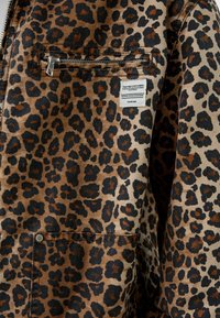 Chaqueta de estampado de leopardo en tonos beige y negro, con un bolsillo en el pecho con cremallera y etiqueta de tela, con costuras visibles y detalles metálicos.