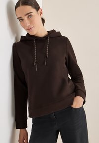 Brązowa bluza z kapturem o textured wykończeniu, z dodatkowymi sznurkami i ściągaczami, zestawiona z ciemnymi dżinsami. Model opiera się o ścianę.