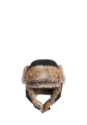 Une femme présente une tenue de sports d’hiver : une veste de ski grise, un pull à motifs, un pantalon et des bottes noirs, un bonnet en fourrure et des lunettes de ski teintées.