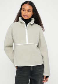 Jasnoszary pulower z polaru z wysokim kołnierzem, półzamek i szerokimi mankietami. Posiada biały pasek na wysokości klatki piersiowej oraz miękką, teksturowaną powierzchnię.