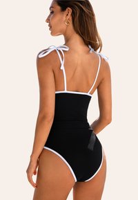 Schwarzer gerippter einteiliger Badeanzug mit weißen Akzenten und Bändern, der ein hochschnittiges Bein-design und eine figurbetonte Silhouette bietet.