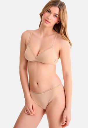 Vrouw met lang haar draagt een bijpassende beige beha en slip met kanten randje, staand tegen een effen lichte achtergrond.