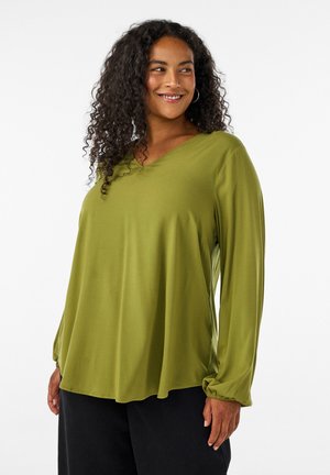 Mulher sorridente com cabelo encaracolado, vestindo uma camisola larga verde-oliva de manga comprida e calças pretas, em pé contra um fundo branco simples.