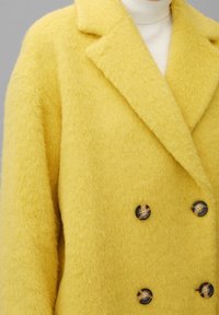 Abrigo de lana amarillo con acabado texturizado, solapas muestradas y frente de doble botonadura con botones marrón oscuro.