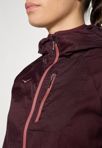 Lähikuva henkilöstä, joka käyttää viininpunaista kevyttä takkia, jossa on vetoketjullinen rintatasku ja siihen sopivat vaaleanpunaiset vetoketjun yksityiskohdat.