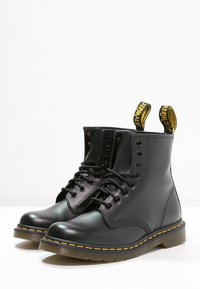 Svarta läderankelbootar med rund tåsko, snörningsdesign, gula sömmar och dragflikar med text på baksidan.