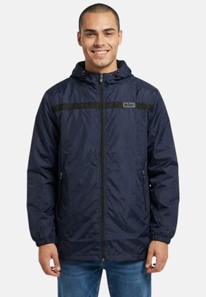 Homme souriant portant un coupe-vent bleu marine à capuche zippée avec une bande horizontale noire et un jean bleu, debout devant un fond blanc.