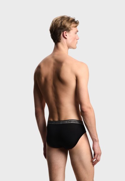 Νέος άνδρας όρθιος χωρίς μπλούζα, με την πλάτη, φορά μαύρα εσώρουχα Emporio Armani, με φόντο απλό ανοιχτό γκρι.