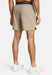 Beige Sportshorts mit einem schwarzen Unter Armour Bund, ausgestattet mit einem durchlöcherten Saumdesign. Getragen mit weißen Sportschuhen.