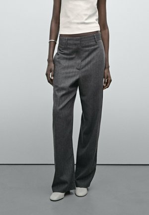 Person trägt hoch taillierte, lockere graue Hose, weißes ärmelloses Oberteil, silbernes Armband und weiße Schuhe und steht vor einem einfarbigen grauen Hintergrund.