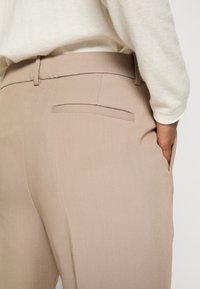 Beige tailored trousers with a smooth texture, featuring a rear welt pocket and a side pocket, paired with a light-colored top.