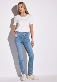 Camiseta blanca de manga corta combinada con jeans de color azul claro, con un corte ajustado, cuatro bolsillos y un diseño sin costuras. Zapatillas blancas completan el look.