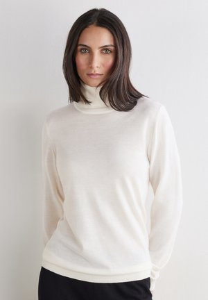 Kobieta o prostych ciemnych włosach, ubrana w biały golf i czarne spodnie na jednolitym jasnym tle.