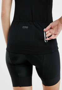 Maglia da ciclismo nera con finitura testurizzata e logo sul retro, abbinata a pantaloni corti neri aderenti e imbottiti. Dettaglio della tasca visibile incluso.