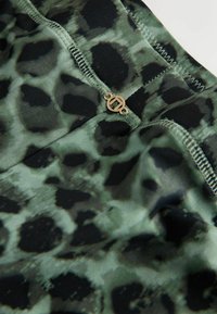 Zaļa leoparda raksta audums ar gludu faktūru, kas rotāts ar melnām vietām, dekoratīvu zelta aparatūru un šuvēm gar jostasvietu.