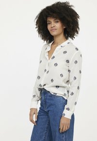 Chemise à manches longues, boutonnée, de couleur beige clair avec un motif floral, fabriquée en tissu doux. Associée à un jean bleu taille haute.