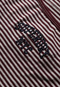 Rot-weiß gestreifter Stoff mit einem bestickten Marine-Design, das "SUPERDRY NYC" liest. Die Textur wirkt weich und lässig.