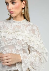 Blouse blanche transparente avec des volants superposés, texture florale, col haut et manches longues. Portée avec de grandes boucles d'oreilles en or.