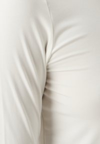 Camicetta fitted a maniche lunghe di un beige chiaro realizzata in un tessuto morbido e elastico con una sottile texture. Dettagli delle cuciture visibili vicino alla spalla.