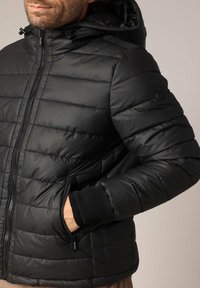 Doudoune noire avec fermeture éclair à l'avant, capuche, design matelassé et poignets côtelés. Comprend des poches latérales et un logo sur la manche.