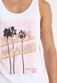 Canotte bianca con una grafica rosa che riporta il testo "RADICAL PARADISE," due palme nere e il nome del marchio "TOM TAILOR DENIM."
