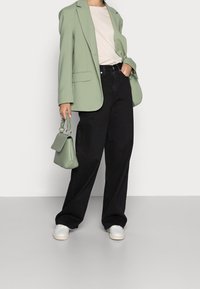 Blazer oversize vert clair sur un top beige, associé à un pantalon large noir. Petit sac à main vert avec une poignée texturée, baskets blanches.