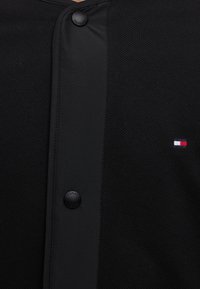 Κοντινή λήψη μαύρου πουκαμίσου Tommy Hilfiger με υφή υφάσματος, δύο μαύρα κουμπιά και μικρό κόκκινο, λευκό και μπλε λογότυπο Tommy Hilfiger στη δεξιά πλευρά.