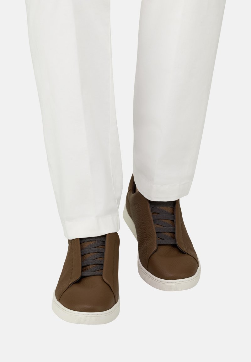 Baskets en cuir marron à semelles blanches portées avec un pantalon blanc, montrant les vues de face et de dessus des chaussures et des jambes inférieures.