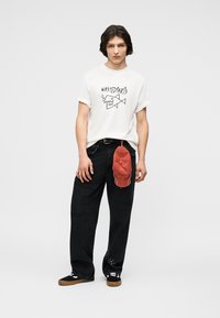 Νεαρός άνδρας που φοράει λευκό γραφικό t-shirt, φαρδιά μαύρα τζιν, μαύρα αθλητικά παπούτσια, με κόκκινο καπέλο να κρέμεται από το λουράκι της ζώνης του.