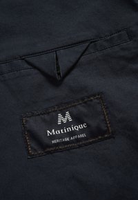 Tessuto blu marino con un dettaglio di design triangolare e un'etichetta tessuta. L'etichetta presenta "Matinique" in bianco con "HERITAGE APPAREL" sottostante.