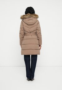 Mujer de pie de espaldas, vistiendo un abrigo acolchado beige de longitud a la rodilla con capucha de piel, cinturón, jeans oscuros y tacones negros, contra un fondo blanco.
