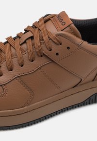 Baskets en cuir marron avec finition texturée, semelle noire contrastante et lacets ronds. Présente un logo sur la languette et des accents cousus.