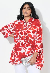 Chemisier blanc avec un grand motif floral rouge, fermeture à boutons sur le devant, manches courtes et coupe fluide ; associé à une pochette argentée.