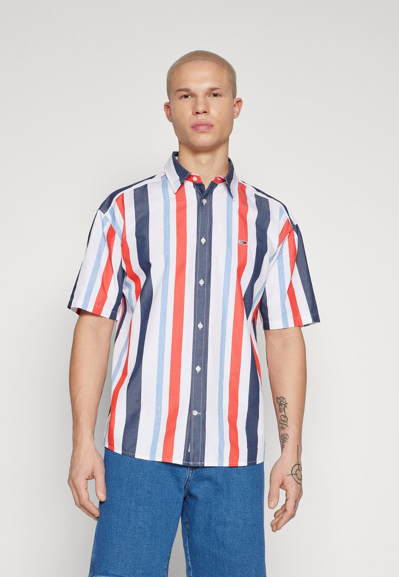 Camicetta a maniche corte con bottoni, caratterizzata da strisce verticali in rosso, blu navy e azzurro. Presenta un colletto e un piccolo logo sul petto.