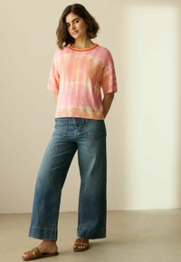 Rosa Batik-Strickpullover mit kurzen Ärmeln und einem kontrastierenden orangen Ausschnitt, kombiniert mit weit geschnittenen Jeans und braunen Sandalen mit offenem Zeh.