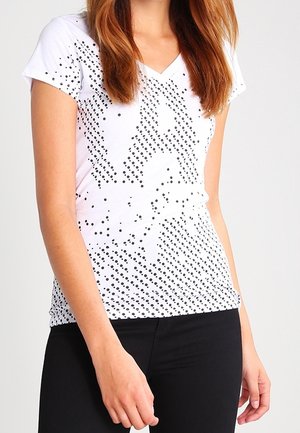 Femme portant un t-shirt blanc à col en V avec un motif diagonal à pois noirs et un pantalon noir, debout devant un fond clair uni.