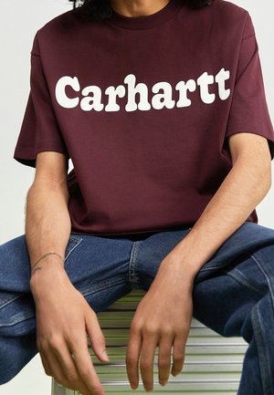 Person in einem kastanienbraunen Carhartt-T-Shirt und blauen Jeans, der mit den Händen auf den Knien sitzt.