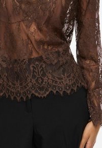 Haut en dentelle marron transparente à manches longues avec des motifs floraux, associé à un pantalon noir, montrant le torse et la main droite sur le côté.