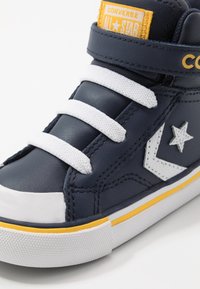 Granatowy sneaker na wysokiej podeszwie z eko skóry, z białymi sznurowadłami i detalami, z żółtą naszywką z logo oraz białym gumowym noskiem i podeszwą.