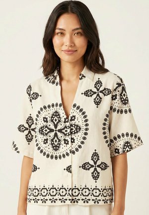Donna che indossa una camicia beige a maniche corte con motivi geometrici e circolari neri, in piedi davanti a uno sfondo chiaro.
