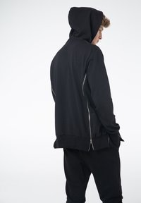 Czarna bluza z kapturem o luźnym kroju, z zamykanymi na zamek kieszeniami po bokach, ściągaczami przy mankietach i dolnym końcu. Gładka tekstura, minimalistyczny design bez wzorów.