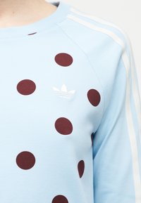 Cămașă de culoare albastru deschis cu mâneci lungi, cu dungi albe pe mâneci, buline mari de culoare roșu închis și logo-ul Adidas Trefoil alb pe piept.