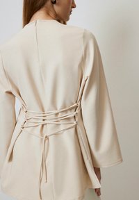 Beige tunic featuring a rounded neckline, long bell sleeves, and crisscross tie details at the back for adjustable fit. Smooth fabric texture.