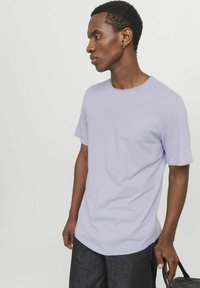 Tricou scurt cu manecă scurtă de culoare violet deschis, cu guler rotund, fabricat dintr-un material moale, purtat cu pantaloni din denim închis și ținând o geantă neagră.